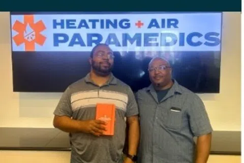 Two men standing in front of a Heating + Air Paramedics sign, one holding an orange box award or gift.