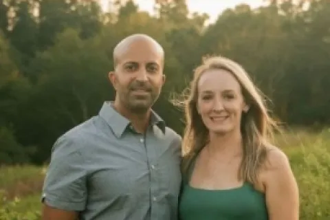 A couple stands together outdoors, smiling in casual attire against a natural backdrop.