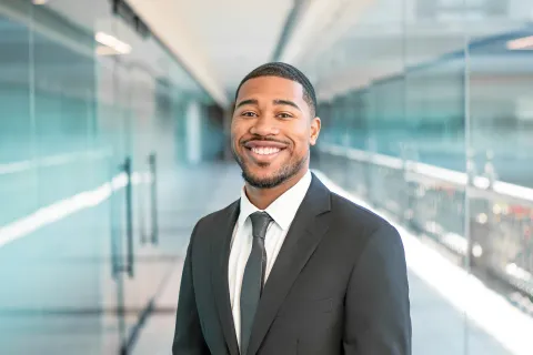 Smiling young businessman wearing a black suit and tie standing in a modern glass office corridor.