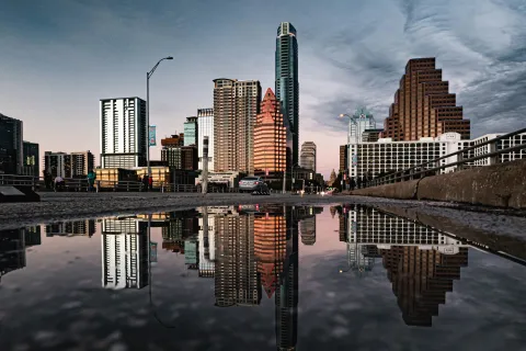City skyline with tall buildings reflected in a large puddle on a street during dusk with cloudy sky.