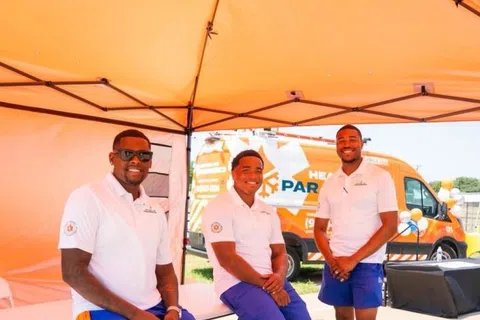 Three men in white shirts and blue shorts under a tent with a Heating Air Paramedics van outside on a sunny day