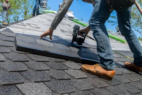 Worker installing asphalt roof shingles on a house using a nail gun on a sunny day.