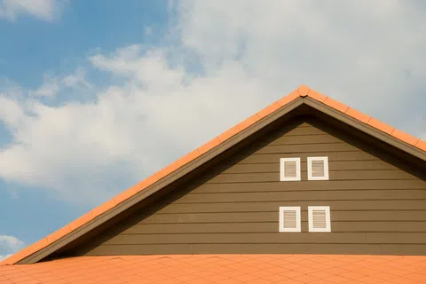 Brown house gable with orange roof tiles and four small white windows under a blue sky with clouds