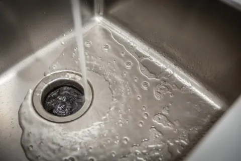 Water flowing into a stainless steel sink drain with bubbles and ripples visible around the drain area.