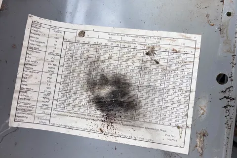 Hand holding a metal panel with a weathered technical specification label outdoors over grass.