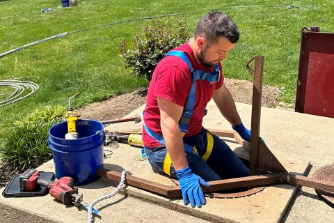 Worker in red shirt and blue gloves opening a manhole cover on a sunny day in a grassy area