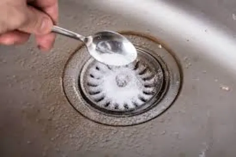 Hand pouring baking soda from a spoon into a kitchen sink drain for cleaning.
