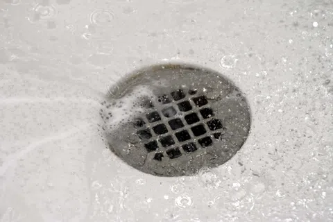 Close-up of a metal sink drain with water and soap bubbles flowing around it in a white basin.