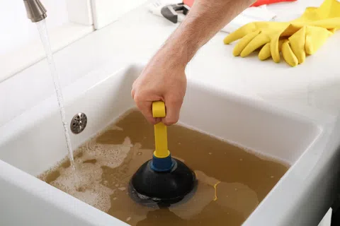 Person using a plunger to unclog a white sink filled with dirty water in a kitchen setting.