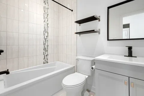 Modern bathroom with white bathtub, toilet, tiled walls, floating shelves, and vanity with black faucet and mirror.