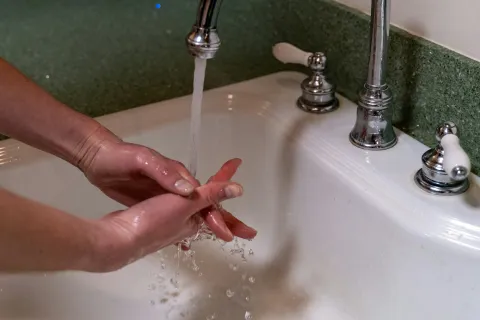 Hands washing under running water from a chrome faucet in a white sink with green backsplash.