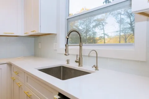Modern white kitchen with quartz countertop, stainless steel sink, and gold hardware beneath a large window.