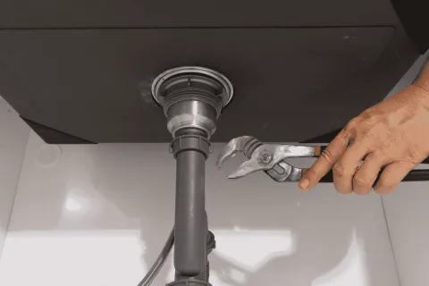 Hand using adjustable wrench to fix plumbing under a kitchen sink with visible pipes and fittings.