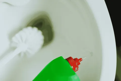 Green bottle with red cap pouring cleaning liquid into a white toilet bowl with brush in background