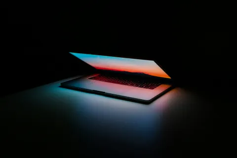 Partially open laptop glowing with colorful screen light in a dark room casting blue and red hues on surface.