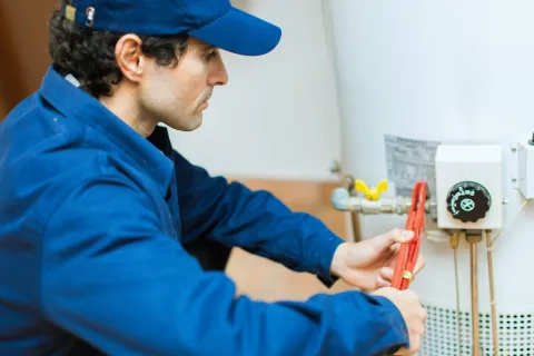 Technician in blue uniform using a wrench to repair a white water heater indoors.