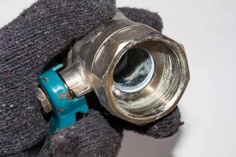 Close-up of a gloved hand holding a metal plumbing valve with a blue handle against a gray background.