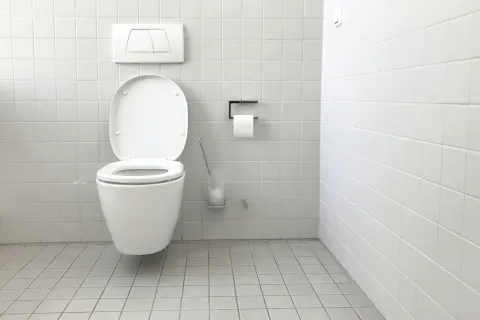 Modern white wall-mounted toilet with open lid in a clean tiled bathroom with toilet paper and brush.