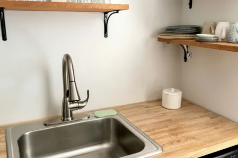 Modern kitchen corner with stainless steel sink, wooden countertop, open shelves with dishes and glasses.