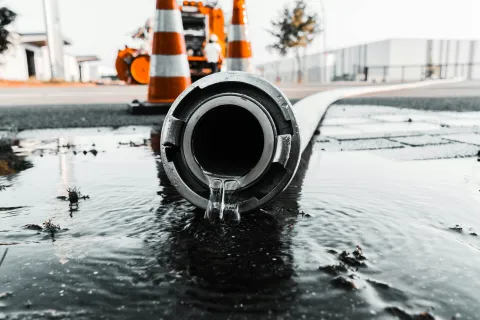 Water flowing from a large drainage pipe onto a wet street with traffic cones and construction equipment in background