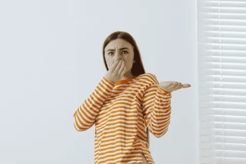 Young woman in orange striped shirt holding nose with disgust against white wall and window blinds