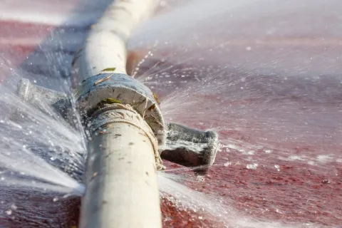 Close-up of a leaking white metal pipe spraying water on a red surface with multiple holes.