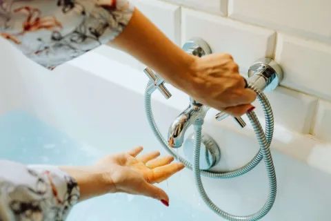 Person testing water temperature from a bathtub faucet with handheld shower in a white tiled bathroom.