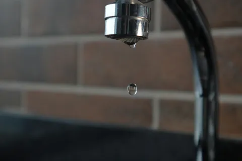 Close-up of a water droplet falling from a kitchen faucet with blurred tiled wall background