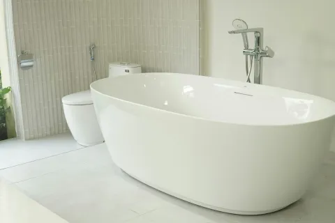 Modern white freestanding bathtub with chrome faucet and toilet in a minimalist bathroom setting.