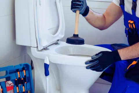 Plumber using a plunger to unclog a white toilet in a bathroom with toolkit nearby.