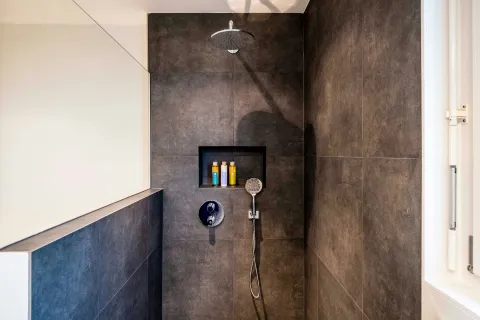 Modern bathroom shower with dark tiles, built-in shelf, overhead rain shower, and handheld showerhead near window.