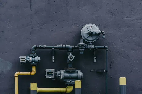 Gas meter and piping setup with yellow and black pipes mounted on a dark gray wall outdoors.