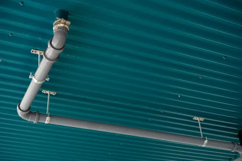Gray metal pipes with joints mounted on a teal corrugated metal ceiling with metal brackets.