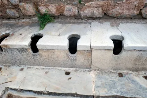 Ancient marble public toilets with multiple stone seats and openings set against a weathered stone wall.
