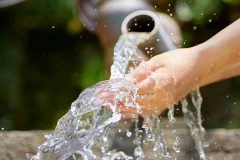 Hands catching clear water flowing from a white pipe outdoors with a blurred natural background