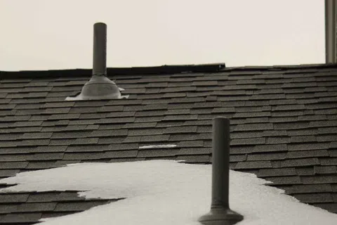 Snow partially covering a dark asphalt shingle roof with two black vent pipes against a white sky background.