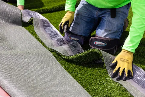 Worker wearing gloves and knee pads installing artificial grass turf outdoors on green surface.