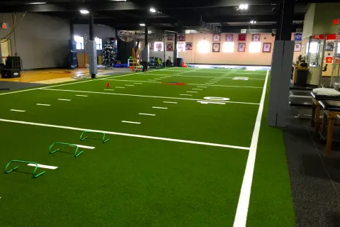 Indoor training facility with artificial turf, agility hurdles, cones, and fitness equipment for sports conditioning.