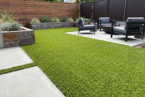 Modern backyard with artificial grass, concrete patio, wooden planters, and black outdoor seating with cushions.