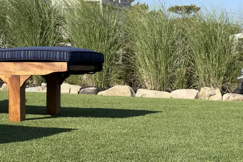 Wooden bench with blue cushion on green artificial grass next to rocks and tall grasses in garden.