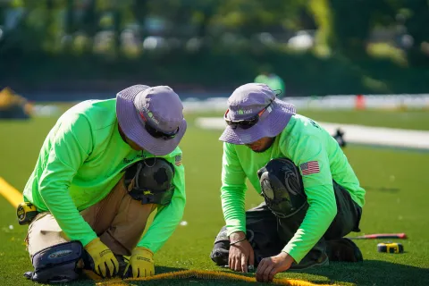 Two workers in bright green shirts and hats installing artificial turf on a sunny day outdoors.
