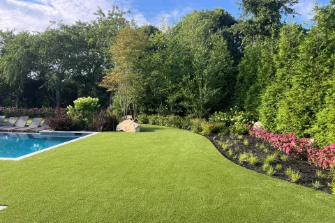 Well-manicured lawn beside a swimming pool with lounge chairs, flower beds, and dense green trees under blue sky.