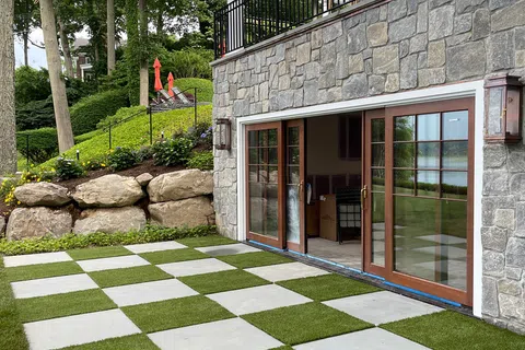 Stone patio with grass tiles in checkerboard pattern outside stone house with large glass doors and greenery
