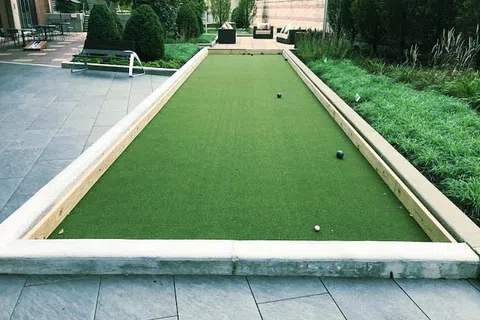 Outdoor bocce ball court with green turf, surrounded by paved patio, bushes, and outdoor seating area.