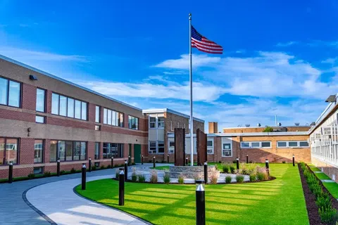 American flag flying on a pole in the courtyard of a modern brick school building under a blue sky.