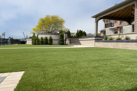 Well-maintained backyard with green lawn, stone retaining walls, trees, and a covered patio attached to a brick house.