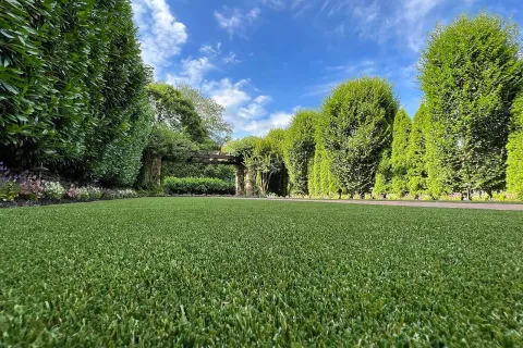 Lush green lawn with neatly trimmed hedges and a wooden pergola under a blue sky with scattered clouds.