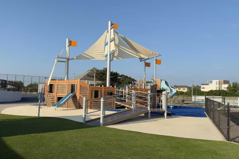 Outdoor playground with ship-themed play structure, slides, climbing ropes, and shade sails under clear blue sky.
