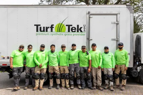 Team of workers in green shirts and knee pads posing in front of Turf Tek USA synthetic grass truck.