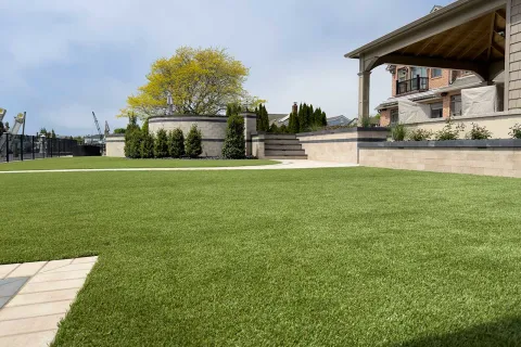 Well-maintained green lawn with modern stone retaining walls, shrubs, and a covered patio in a residential backyard.